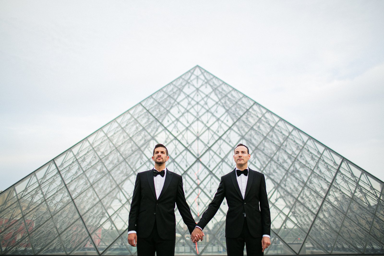 Pareja de novios de dos hombres cogidos de la mano con parte de una pirámide del museo Louvre detrás