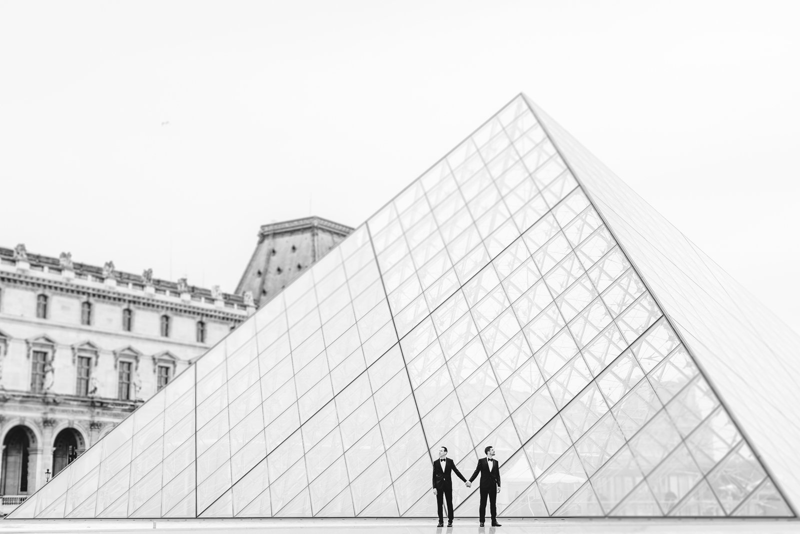 Pareja de novios de dos hombres cogidos de la mano con una pirámide del museo Louvre detrás. Fotografía en blanco y negro.