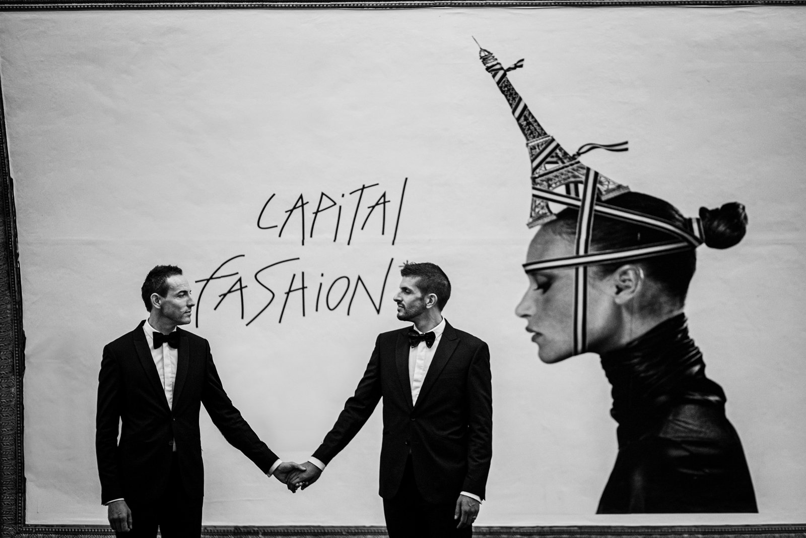 Pareja de novios de dos hombres cogidos de la mano con una ilustración detrás de una mujer con la torre Eiffel de sombrero y la inscripción Capital Fashion. Fotografía en blanco y negro.