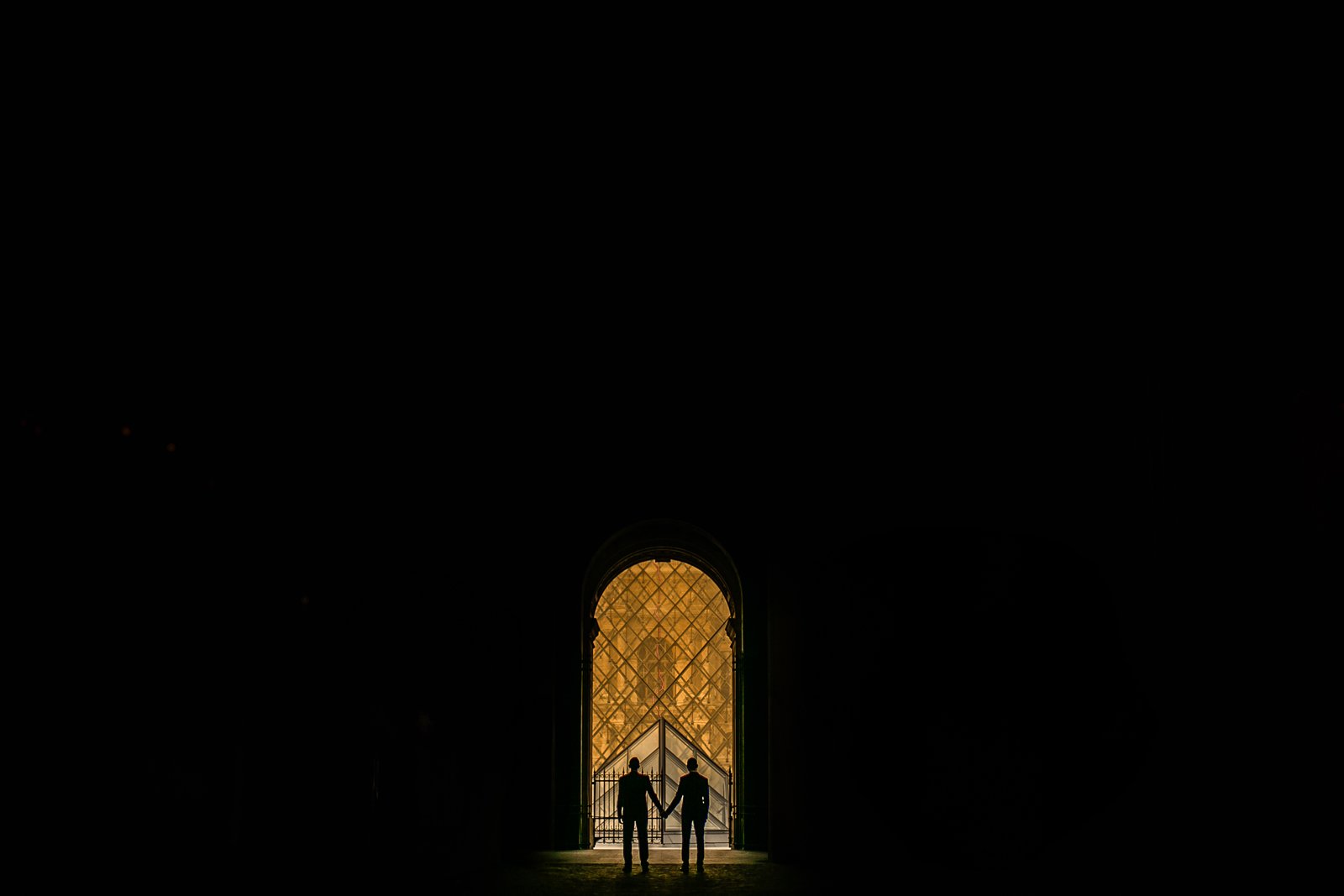Pareja de novios de dos hombres cogidos de la mano bajo un gran portón antiguo, a contraluz