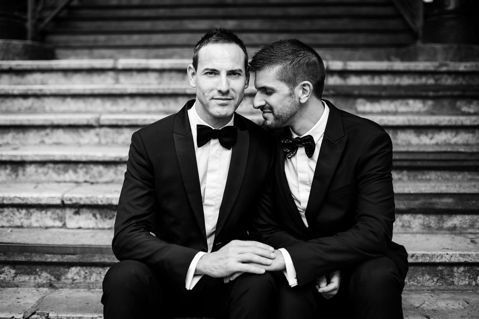 Pareja de novios de dos hombres sentados en una escalera, cogidos de la mano y en actitud romántica. Fotografía en blanco y negro.