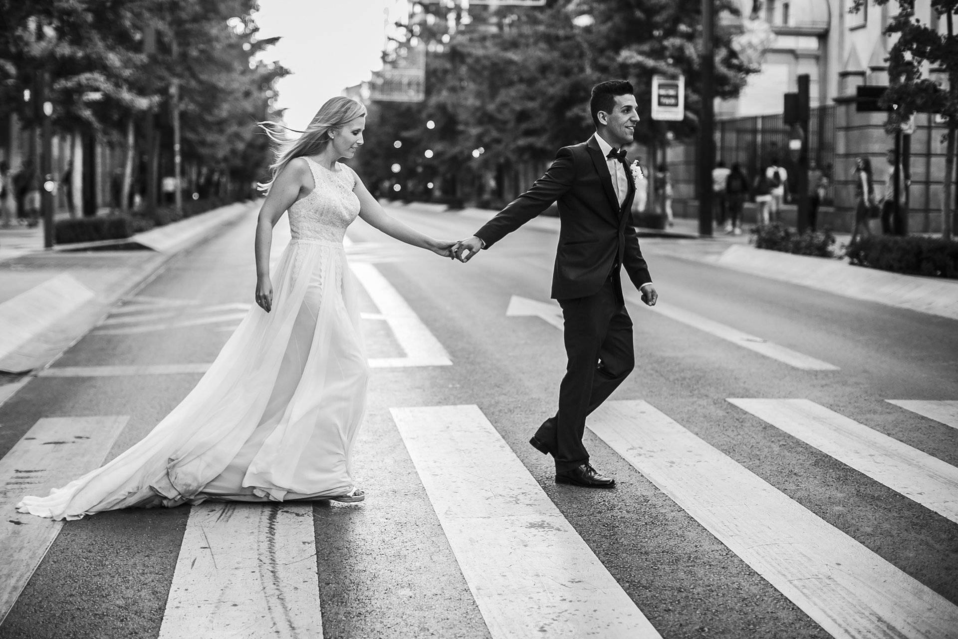 Fotografía en blanco y negro de pareja de novios con vestido blanco y esmoking curzando un paso de cebra