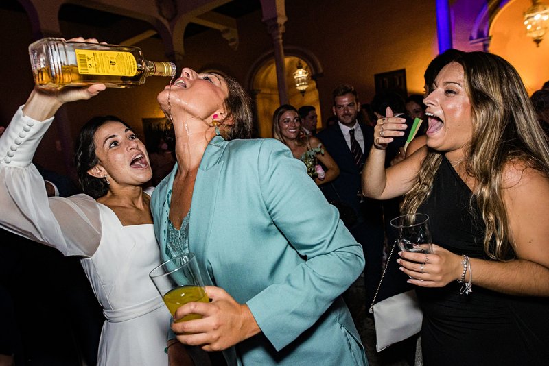 Novia con vestido blanco de fiesta con dos amigas, dándoles de beber directamente de la botella
