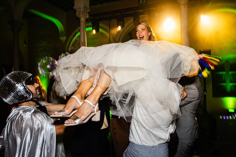 Novia vestida de blanco llevada en volandas por los invitados de la boda durante el baile, mientras pone cara divertida