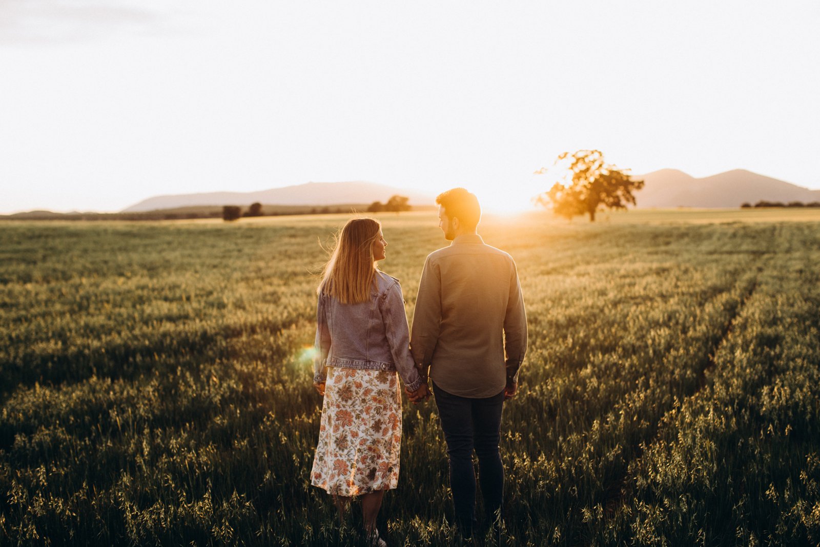 Pareja cogidos de la mano mirándose mutuamente, mientras se pone el sol, en medio de un prado con algunos árboles