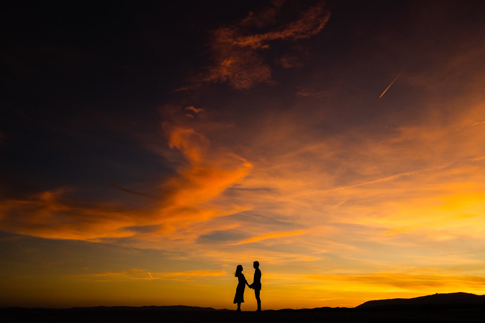 Sesión de fotos en pareja en Granada: pareja cogida de las manos, mirándose de frente, en un atardecer con nubes en el campo