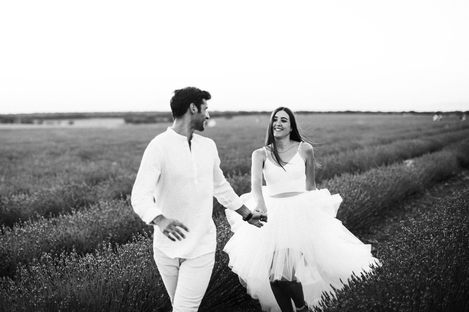 Fotografía en blanco y negro: pareja corriendo por mitad de un campo de lavanda, ambos vestidos de blanco, cogidos de la mano, él delante y ella detrás, el vuelto hacia atrás para mirarla. Ambos sonríen.