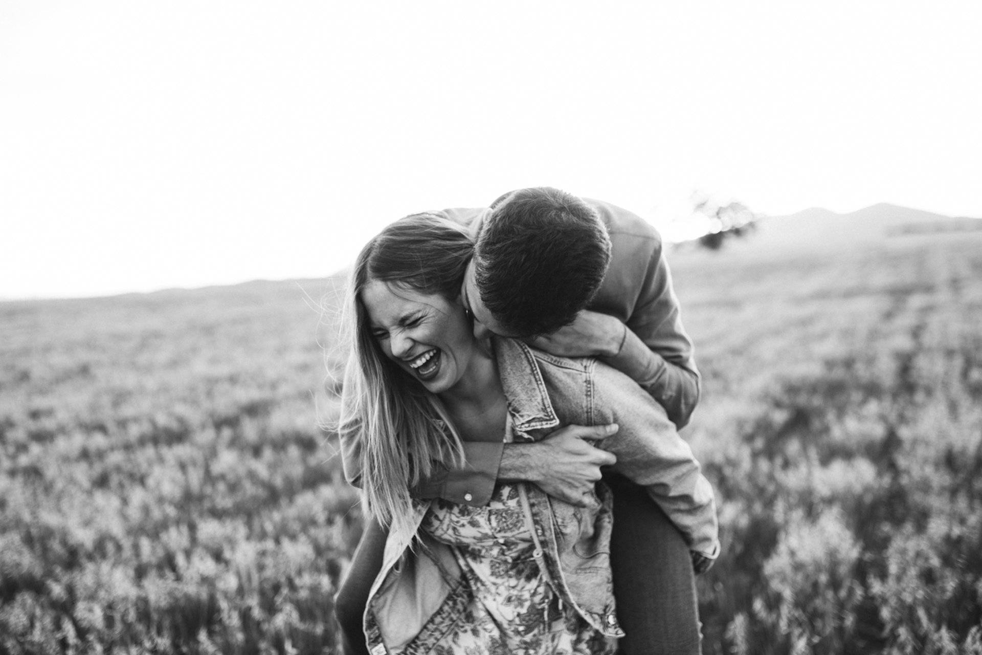 Fotografía en blanco y negro de pareja en un prado, en la que ella lo carga a él a caballito mientras ambos ríen