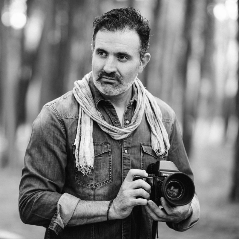 Alfonso en el bosque sostiene una cámara de fotos profesional en sus manos mientras mira lateralmente. Fotografía en blanco y negro.