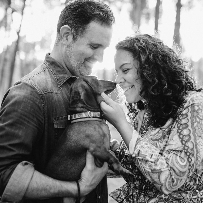 Plano medio de Alfonso y Ana en el bosque, ambos sonriendo mientras juegan con su perrito, sostenido en brazos por Alfonso. Fotografía en blanco y negro