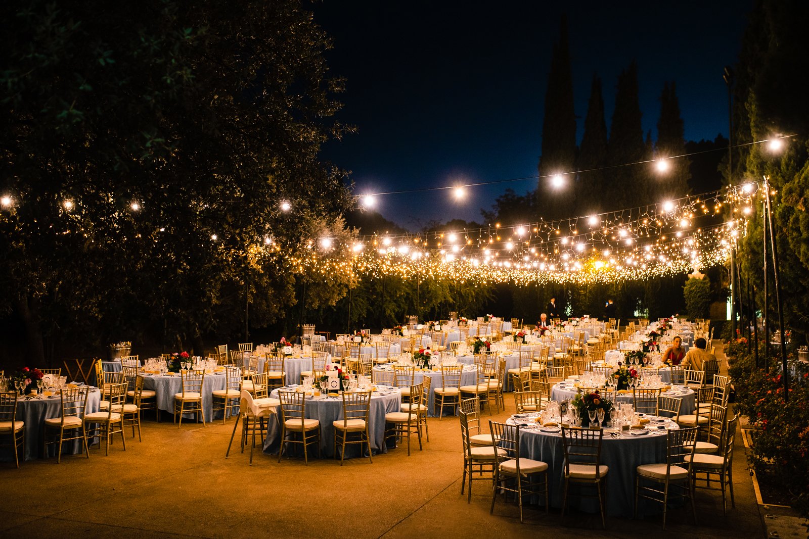 Vista de muchas mesas redondas preparadas para la celebración de na boda, con centros de flores y todos los detalles, iluminadas por muchas bombillas, de noche y al aire libre