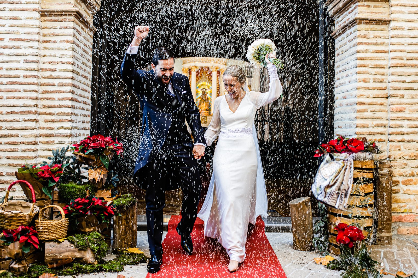 Pareja de novios saliendo de la iglesia sobre una alfombra roja, con flores y decoración a ambos lados, mietras tratan de protegerse de la lluvia de arroz que le están lanzando