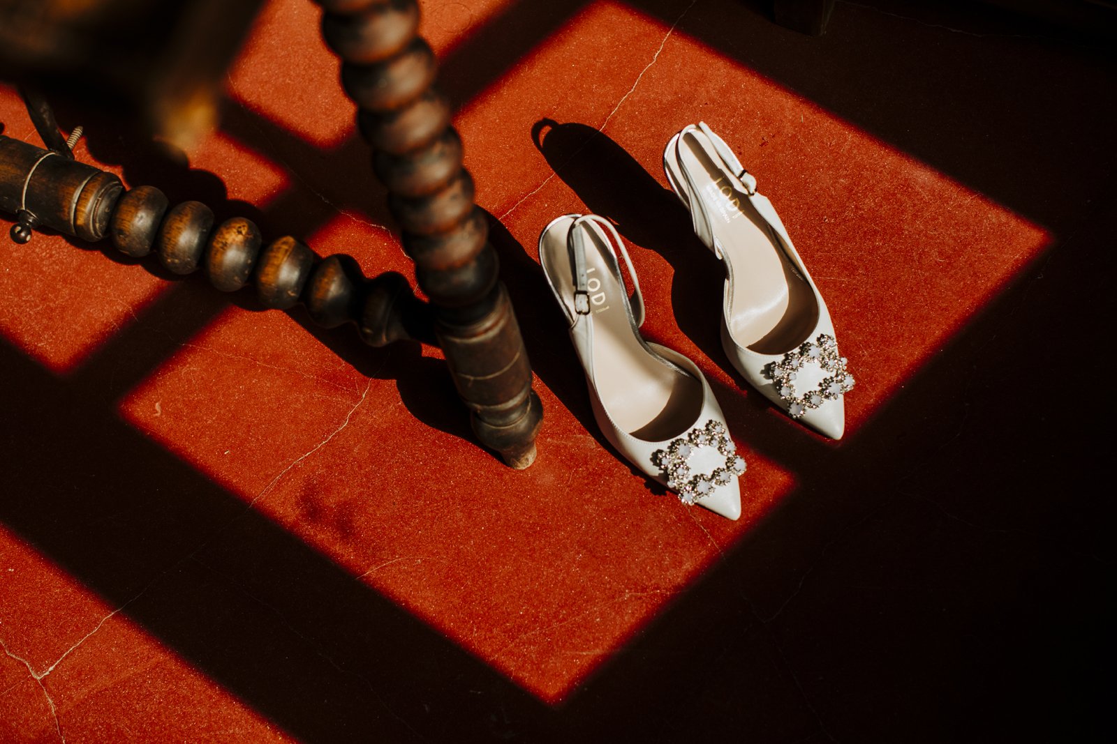Zapatos de novia sobre suelo rojo, junto a las patas de algún mueble rústico