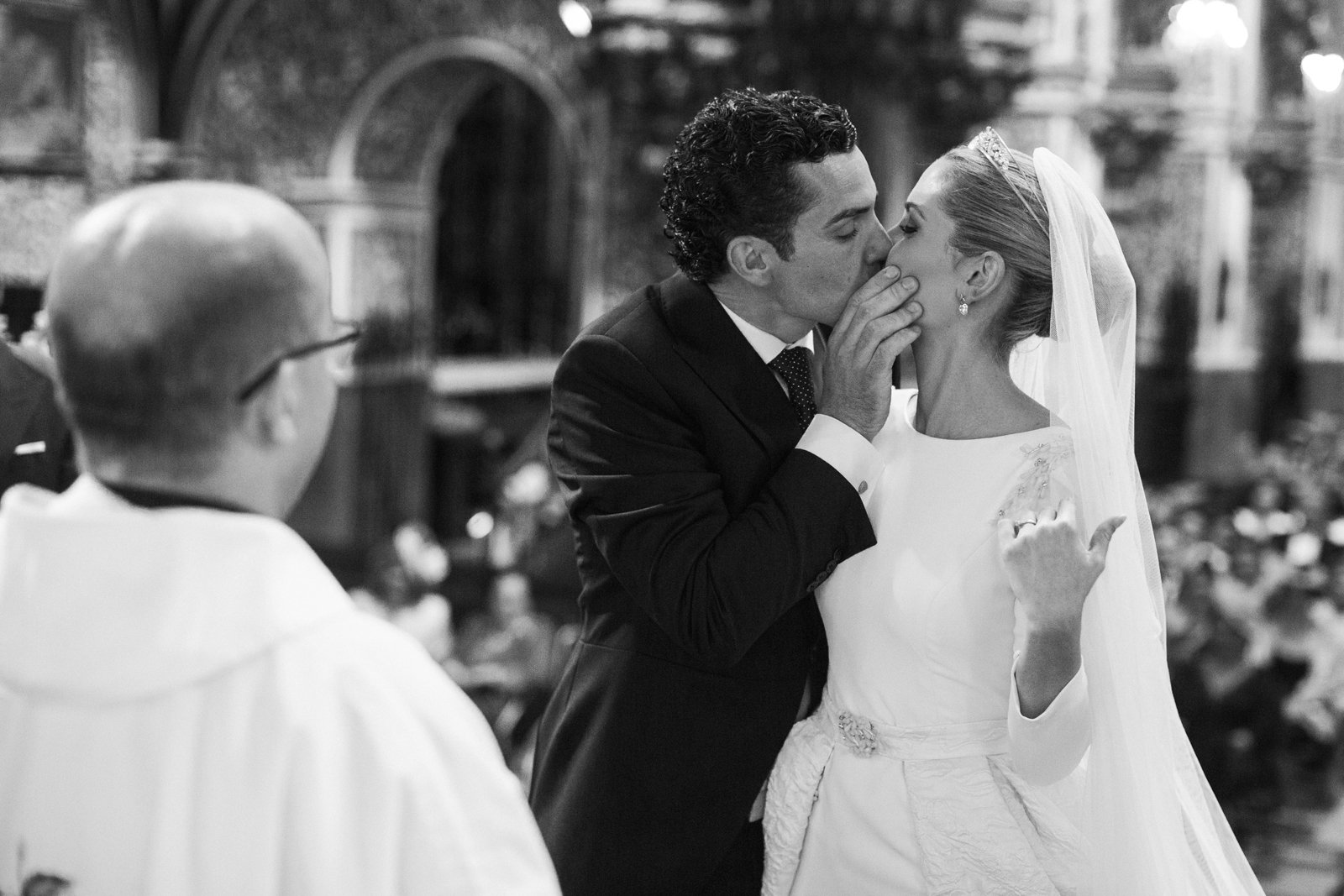 Pareja de novios en el altar de la iglesia, el novio besa a la novia mientras el cura mira