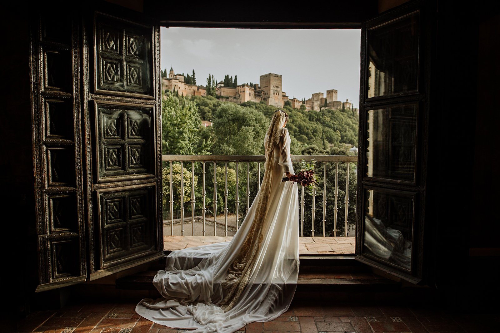 Novia vestida de blanco asomada a un gran balcón antiguo, desde el que se ve una hermosa vista de la Alhambra de Granada