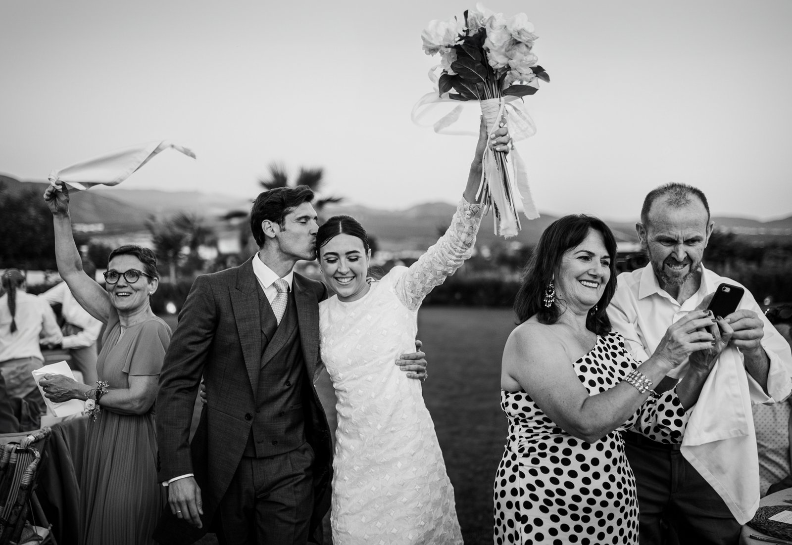 Novios en la celebración de su boda al aire libre. La novia levanta el ramo y el novio la coge por la cintura mientras una pareja de invitados miran el movil y otra invitada blande un pañuelo