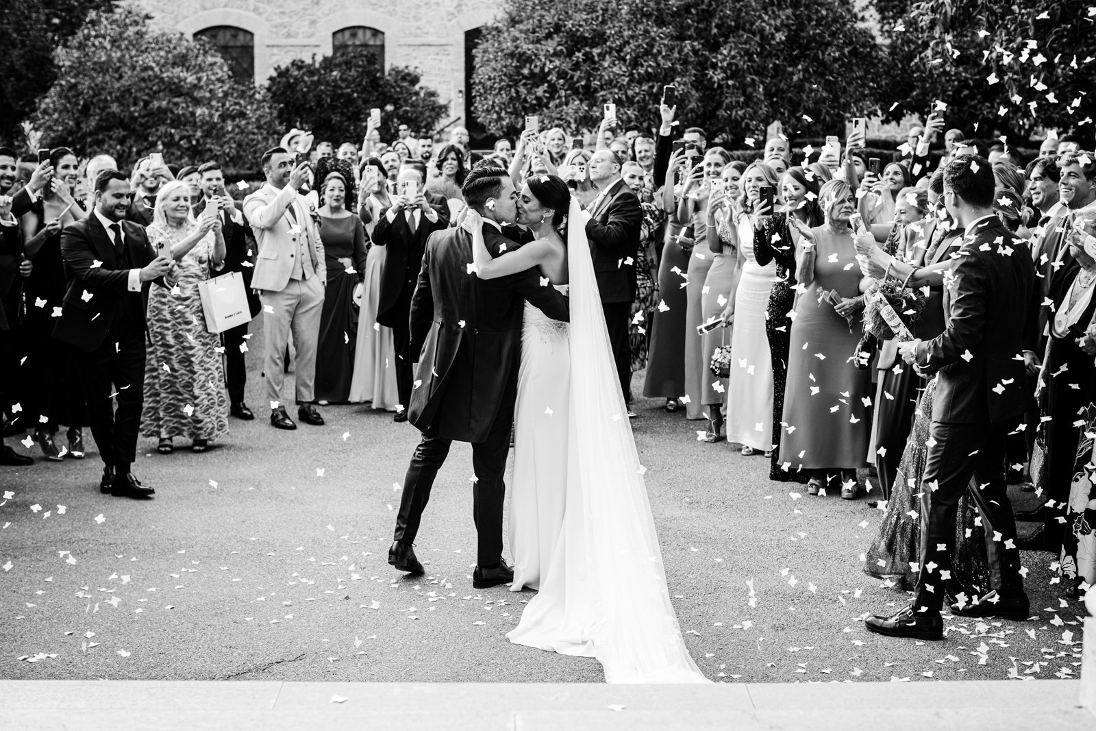 Novios de pie besándose rodeados por todos los invitados, lanzándoles confeti y grabándolos con el móvil