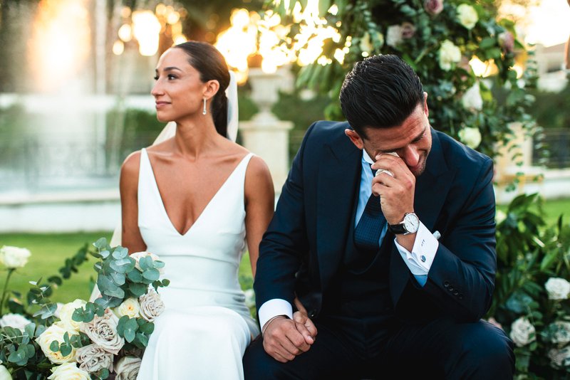 Pareja de novios en un jardín con rosas. Mientras ella mira lateralmente, él se limpia el ojo con un pañuelo.