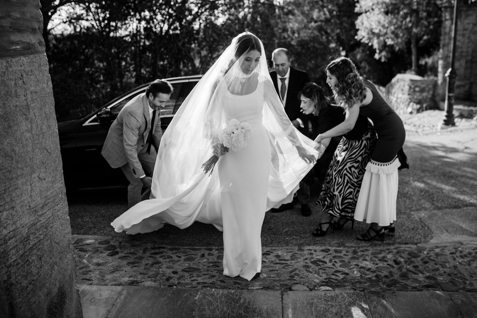 Fotografía en blanco y negro de novia con vestido blanco bajando del coche y familiares colocándole la cola del vestido